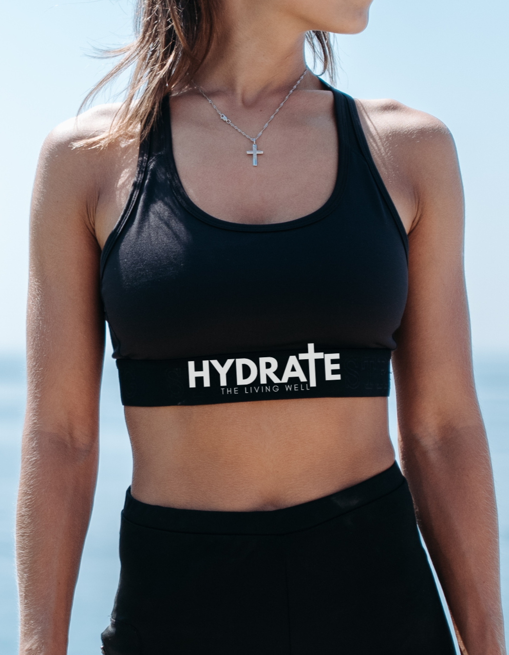 Person wearing black sports bra and leggings with 'HYDRATE' branding by the ocean.
